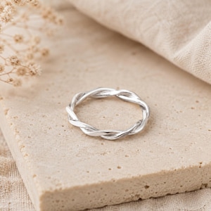 Può includere: Un anello in argento lucido caratterizzato da un design intrecciato simile a una corda. La fascia è composta da due fili di metallo intrecciati che creano un motivo circolare continuo. La superficie dell'anello è liscia e riflettente.
