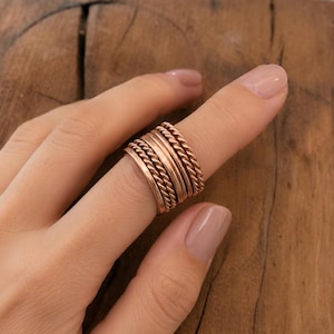 Puede incluir: Un conjunto de anillos apilables con múltiples bandas en un tono cobre cálido. El diseño consiste en bandas planas lisas y bandas texturizadas con estilo de cuerda trenzada, creando una apariencia de capas al usarlas juntas en un dedo.