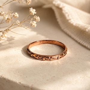 May include: A copper-toned metal ring featuring a continuous embossed scroll pattern around the exterior band. The ring has a warm, metallic finish with subtle dark oxidation within the recessed areas of the intricate, repeating swirl design.