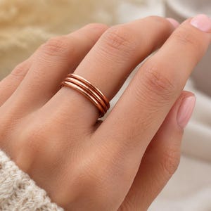 Op de afbeelding: Een gepolijste roségouden ring met een ontwerp van drie banden, bestaande uit drie gladde, afgeronde en met elkaar verbonden cirkelvormige draden. Het metaal heeft een warme, koperkleurige afwerking die het licht gelijkmatig weerkaatst.