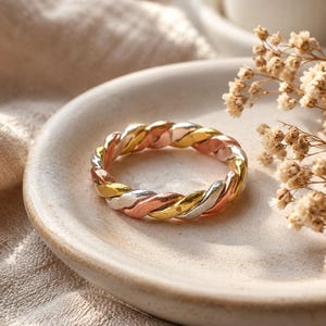 May include: A tri-colour ring featuring a twisted design with alternating strands of yellow gold, rose gold, and silver-toned metal. The band is constructed from three distinct metallic wires woven together to create a continuous braided pattern.