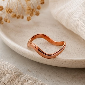May include: A polished copper-toned ring featuring a continuous, smooth, undulating wave-like design. The metal band has a reflective, metallic finish with a consistent thickness throughout its curved, flowing shape, creating a simple, singular loop.