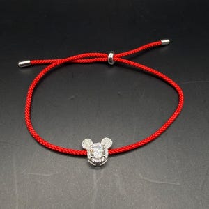 May include: Red cord bracelet featuring a silver-toned charm in the shape of a mouse head, encrusted with clear crystals. The bracelet has adjustable ends with silver-toned metal tips and a sliding knot closure. The background is black.