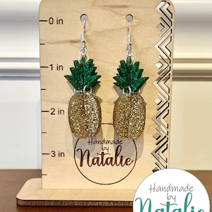 May include: Gold glitter pineapple earrings with green glitter leaves and silver hooks. The earrings are displayed on a wooden stand with a ruler measuring up to 3 inches. The stand also has a circular label that says "Handmade by Natalie".