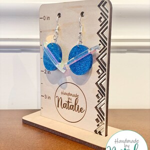 May include: A pair of blue glitter earrings with iridescent accents on a wooden display stand. The earrings are approximately 2 inches long. The stand has a ruler on the side with measurements in inches. The stand also has the text "Handmade by Natalie" on it.