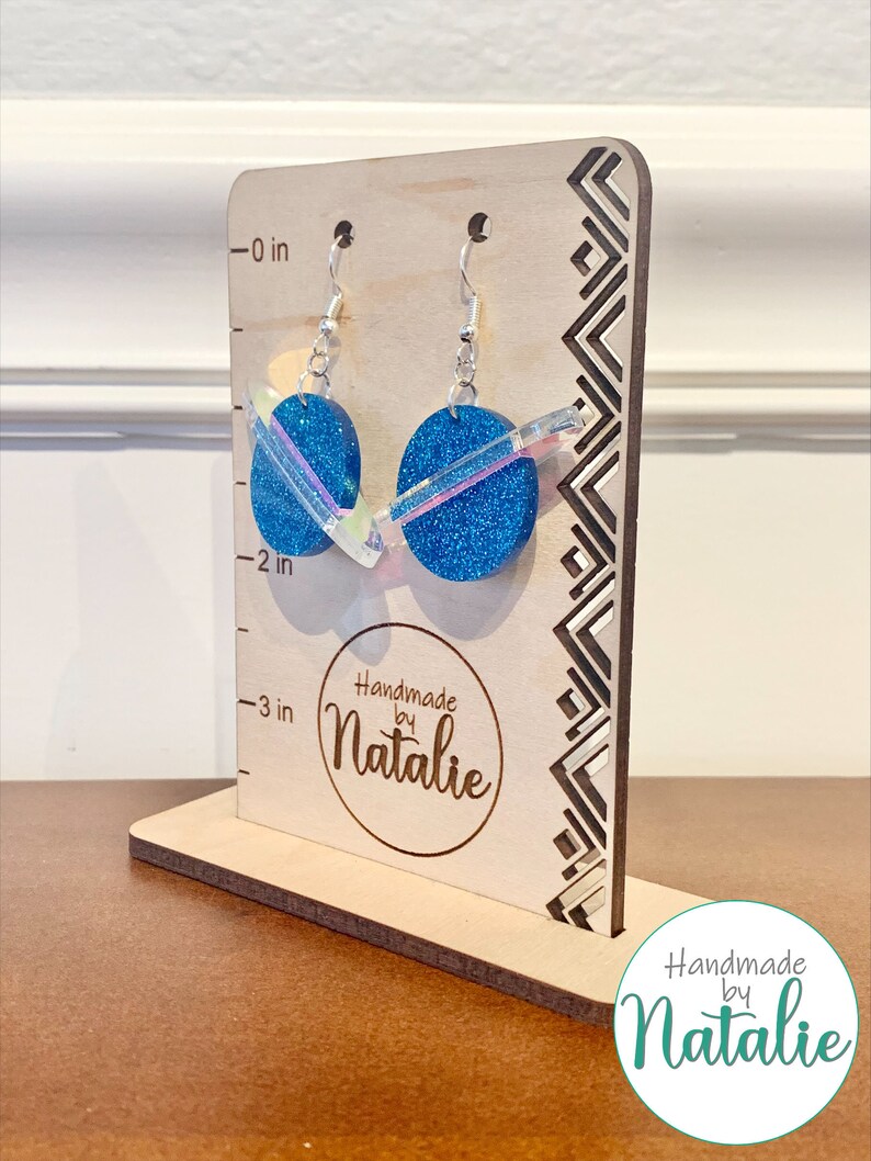 May include: A pair of blue glitter earrings with iridescent accents on a wooden display stand. The earrings are approximately 2 inches long. The stand has a ruler on the side with measurements in inches. The stand also has the text "Handmade by Natalie" on it.