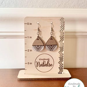 Op de afbeelding: Een paar druppelvormige houten oorbellen met een geometrisch patroon, gepresenteerd op een lichthouten standaard met een liniaal in inches. De standaard bevat de tekst Handmade by Natalie en een verticaal decoratief randpatroon.