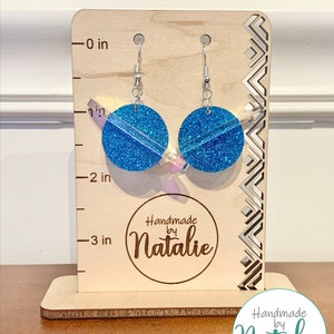 May include: A pair of blue glitter earrings with silver hooks, measuring approximately 2 inches in length. The earrings are displayed on a wooden stand with a "Handmade by Natalie" logo.