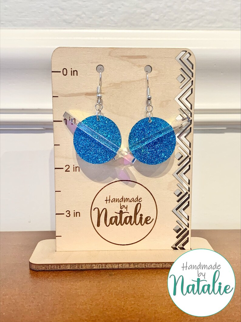 May include: A pair of blue glitter earrings with silver hooks, measuring approximately 2 inches in length. The earrings are displayed on a wooden stand with a "Handmade by Natalie" logo.
