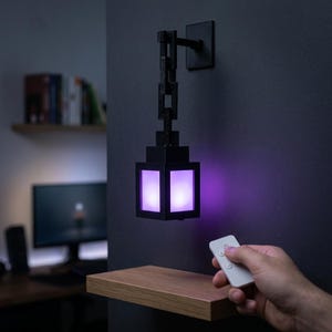 Pode incluir: Uma lâmpada de parede preta com um design pixelizado, emitindo um brilho roxo suave. A lâmpada é suspensa por uma corrente e montada em uma parede escura. Um controle remoto é segurado na mão, presumivelmente para controlar a lâmpada.