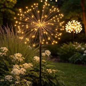 May include: Outdoor decorative light fixtures shaped like starbursts. Each fixture has numerous small, warm-toned LED lights radiating from a central point. The lights are mounted on black poles, set in a garden with greenery and white flowers.