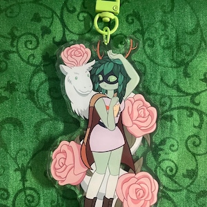 May include: A clear acrylic keyring featuring a cartoon character with green skin, a mask, and antlers, surrounded by pink roses and a white animal. The keyring has a lime green clasp.
