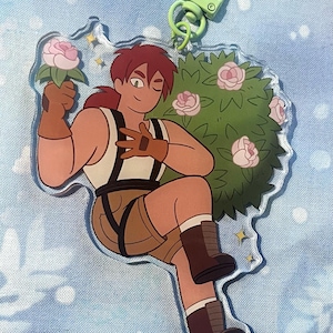 May include: A clear acrylic charm featuring a cartoon character with brown hair, wearing a white tank top, brown shorts, and boots. The character is holding a pink flower and is next to a green bush with pink flowers. A green clasp is attached.