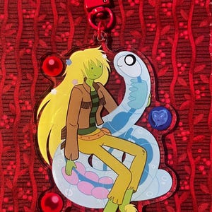 May include: A clear acrylic charm featuring a cartoon character with long blonde hair, a brown jacket, and yellow pants, seated on a light blue snake. The charm has a red clasp and decorative red and blue accents.