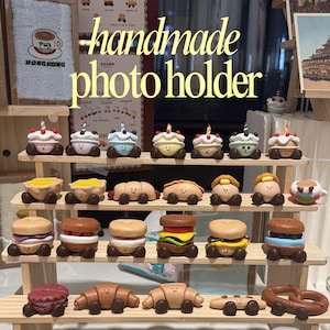 May include: A wooden display shelf filled with handmade photo holders shaped like miniature desserts. The photo holders include cupcakes, sandwiches, croissants, donuts, and pretzels, all with smiling faces. The text "handmade photo holder" is visible.