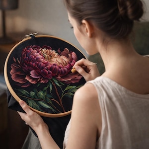Puede incluir: Un bastidor de bordado circular de madera que sostiene un panel de tela negra con un diseño detallado de una gran flor de peonía de múltiples capas en tonos magenta intenso, morado y melocotón suave, con hojas verdes.