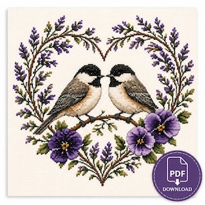 May include: Cross-stitch pattern featuring two birds perched on a branch within a heart-shaped wreath of purple flowers and greenery. The birds have black caps and gray and brown bodies. A PDF download icon is in the lower right corner.