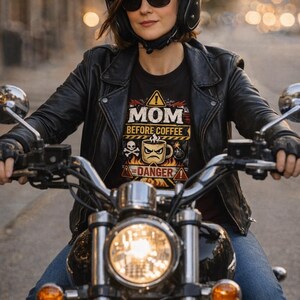 May include: A woman wearing a black motorcycle helmet and sunglasses, seated on a motorcycle. She wears a black leather jacket and a black t-shirt with the text "MOM BEFORE COFFEE" and a coffee cup graphic. The motorcycle has a bright headlight.
