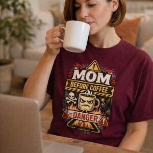 May include: A maroon t-shirt with the text "MOM BEFORE COFFEE" and a graphic of a coffee cup with a skull and crossbones. A woman is drinking from a white mug while working on a laptop.