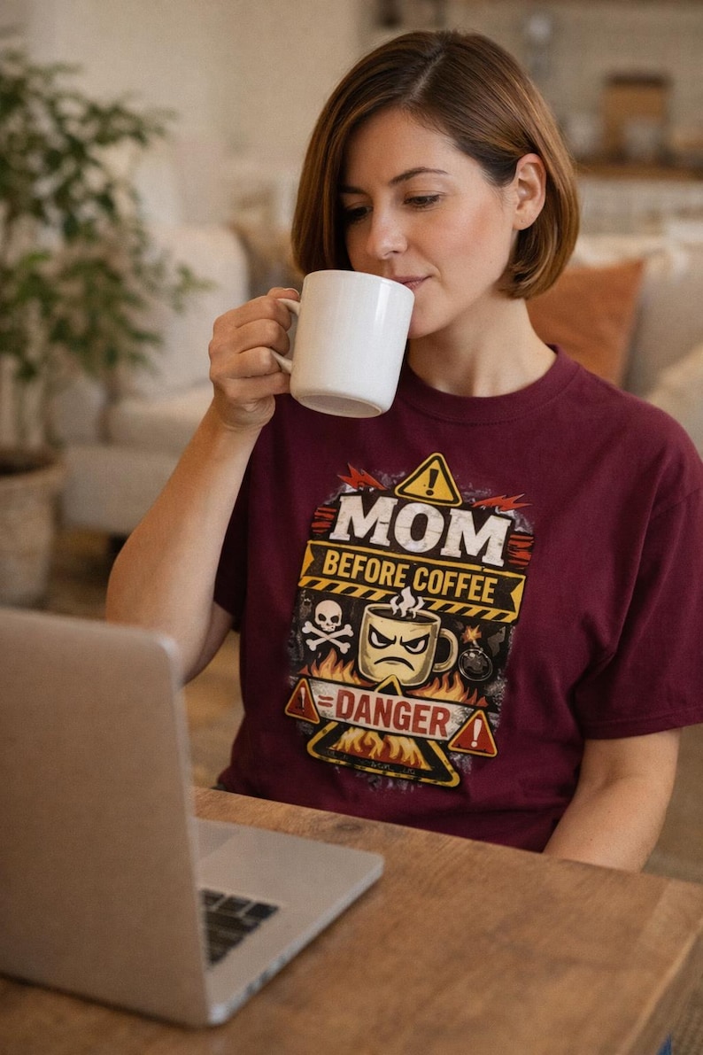 May include: A maroon t-shirt with the text "MOM BEFORE COFFEE" and a graphic of a coffee cup with a skull and crossbones. A woman is drinking from a white mug while working on a laptop.
