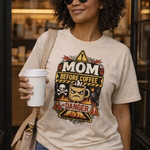 May include: A woman wearing a beige t-shirt with the text "MOM BEFORE COFFEE" and a graphic of an angry coffee cup, skull, and bomb. She is also wearing blue jeans and sunglasses, and holding a white coffee cup.