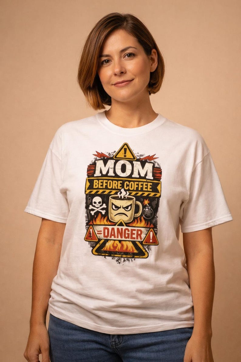 May include: White t-shirt with a humorous graphic. The design features the text "MOM BEFORE COFFEE" above a grumpy coffee cup, skull, and bomb, with "DANGER" below. The shirt is worn by a person.