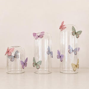 May include: Three clear glass cylindrical vases of varying heights, each decorated with multiple colorful butterfly embellishments in shades of pink, purple, blue, and yellow, attached to the exterior surfaces of the glass containers.