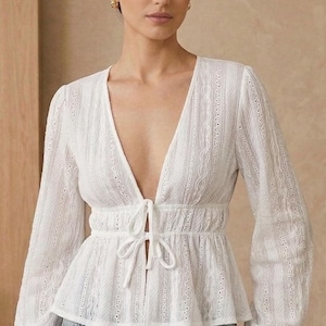 Può includere: Camicetta bianca a maniche lunghe con scollo a V profondo e chiusura con fiocco. La camicetta presenta intricati dettagli traforati e vita stretta. Le maniche hanno polsini svasati e arricciati. La modella indossa jeans blu.