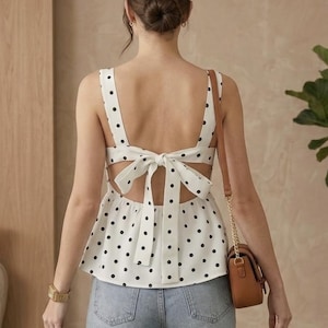 May include: White sleeveless top with black polka dots, featuring a bow detail at the back. The top has a peplum hem and wide straps. The model is wearing light blue jeans and a brown purse with a gold chain.