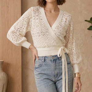 Peut inclure: Un pull cache-cœur crème avec un col en V et des manches longues à motifs. Le pull a une fermeture à nouer à la taille et est associé à un jean bleu clair. Une montre en or est visible au poignet.