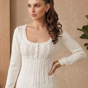 Peut inclure: Pull-over crème avec un col rond et des manches longues. Le pull présente un motif en tricot torsadé sur la partie inférieure et un motif texturé sur le haut de la poitrine, avec trois boutons. Une montre dorée est visible au poignet.
