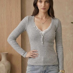 Op de afbeelding: Een grijze geribbelde top met lange mouwen en een V-halslijn afgezet met wit kant. De top heeft een knoopsluiting en wordt gecombineerd met een lichtblauwe jeans. Een gouden horloge en sieraden zijn zichtbaar.