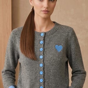 May include: A grey cardigan featuring a blue heart motif and matching blue buttons. The sleeves have light blue ribbed cuffs. This button-down sweater is a stylish addition to any wardrobe.