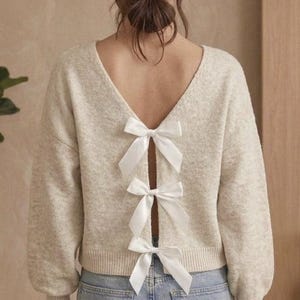Peut inclure: Un pull beige à col en V et manches longues. Le dos du pull est orné de trois nœuds en satin blanc. Le modèle porte un jean bleu clair et une montre dorée. Ce pull est un ajout élégant à toute garde-robe.