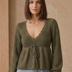 Peut inclure: Pull en tricot vert olive avec un décolleté en V festonné et des manches longues. Le pull a un détail de cordon de serrage à la taille et est associé à un jean bleu. Un collier et une montre en or complètent le look.