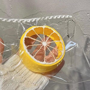 May include: A yellow lemon slice keyring with a white rind and a clear centre. The keyring is encased in a clear plastic bag and has a silver chain attached.