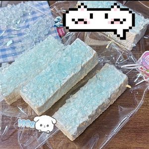 May include: Several rectangular, light blue, and white treats are visible, wrapped in clear plastic. The treats have a textured, crystalline appearance. A cartoon cat face is in the upper right corner. The treats are packaged in a clear plastic bag.