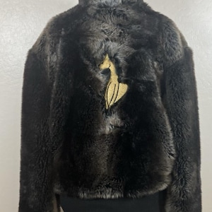 May include: A dark brown faux fur jacket with a high collar and a gold-colored emblem on the back. The jacket has a black ribbed hem and cuffs. The jacket is displayed on a white mannequin.