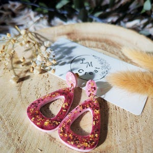 May include: A pair of pink and burgundy resin earrings with gold glitter accents. The earrings have a teardrop shape with a hollow center and are displayed on a wooden surface. The earrings have a matching pink stud.