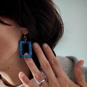 May include: Blue rectangular earrings with gold-coloured hooks. The earrings have a glittery, speckled appearance. The image shows the earrings being worn, with a hand touching one of them. The earrings are a unique accessory.