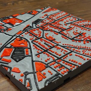 May include: A 3D-printed map with a gray base, black roads, and red buildings. The raised relief map shows a detailed urban layout, with various structures and street patterns. The map is square and sits on a wooden surface.