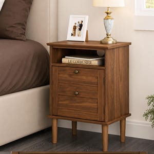 May include: A brown wooden nightstand with an open storage shelf and two drawers featuring small round knobs. The nightstand stands on four tapered legs and includes a book titled LE CORBUSIER on the shelf, a picture frame, and a table lamp.
