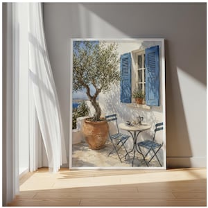 May include: A framed wall art print depicting a Mediterranean terrace scene with a potted olive tree, two blue metal folding chairs, and a small round table. The scene features a white wall with open blue shutters, a window, and a view of the sea.