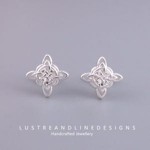 Può includere: Un paio di orecchini a lobo color argento con un motivo a nodo celtico. Ogni orecchino presenta un design a forma di quadrifoglio. Gli orecchini sono esposti su uno sfondo grigio chiaro. Il testo "LUSTREANDLINEDESIGNS Handcrafted Jewellery" è in basso.