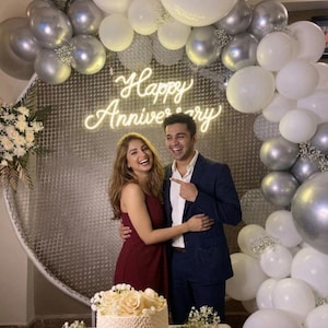 以下が含まれることがあります： 「Happy Anniversary」と書かれたネオンサインと、銀と白の風船を背景に笑顔のカップル。女性はバーガンディ色のドレス、男性はネイビーのスーツを着ています。白いフロスティングと花で飾られたケーキが前景にあります。