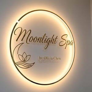 Può includere: Un'insegna circolare con le parole "Moonlight Spa" in elegante scrittura dorata, illuminata da una luce calda e brillante. Sotto, è visualizzato il testo "by Olivia Chen". L'insegna presenta anche un design a fiore di loto e onde.