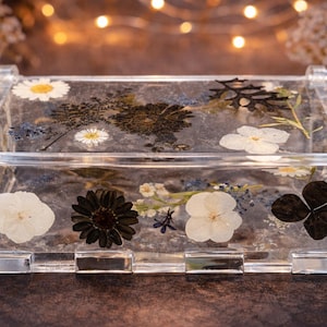 Puede incluir: Caja transparente de acrílico con flores secas incrustadas, incluyendo margaritas blancas, flores marrón oscuro y pétalos blancos. La caja tiene forma rectangular con tapa con bisagras y está diseñada para exhibir la belleza natural de los elementos botánicos preservados.