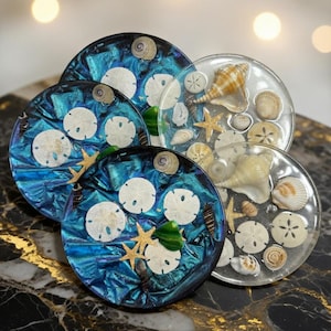 Puede incluir: Un conjunto de cinco posavasos redondos. Los posavasos presentan conchas marinas, estrellas de mar y dólares de arena incrustados en resina transparente. Algunos posavasos tienen un fondo azul iridiscente, otros son transparentes.
