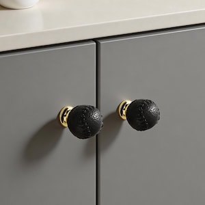 May include: Two black cabinet knobs with a textured surface, resembling a baseball, are mounted on a gray cabinet. The knobs have a gold-colored base. A small white pot with pink flowers sits on the countertop above.