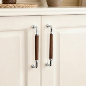 May include: Two cabinet pulls with a polished chrome finish and a dark wood centre. The pulls are mounted on a white cabinet door. The design is a modern take on a classic style, suitable for kitchen or bathroom cabinetry.